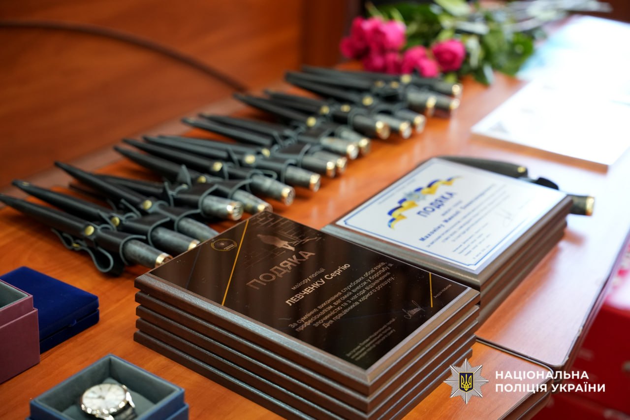 Award plaques and a row of ceremonial pens on a wooden table, with pink flowers in the background and a Ukrainian Police emblem in the bottom right corner.