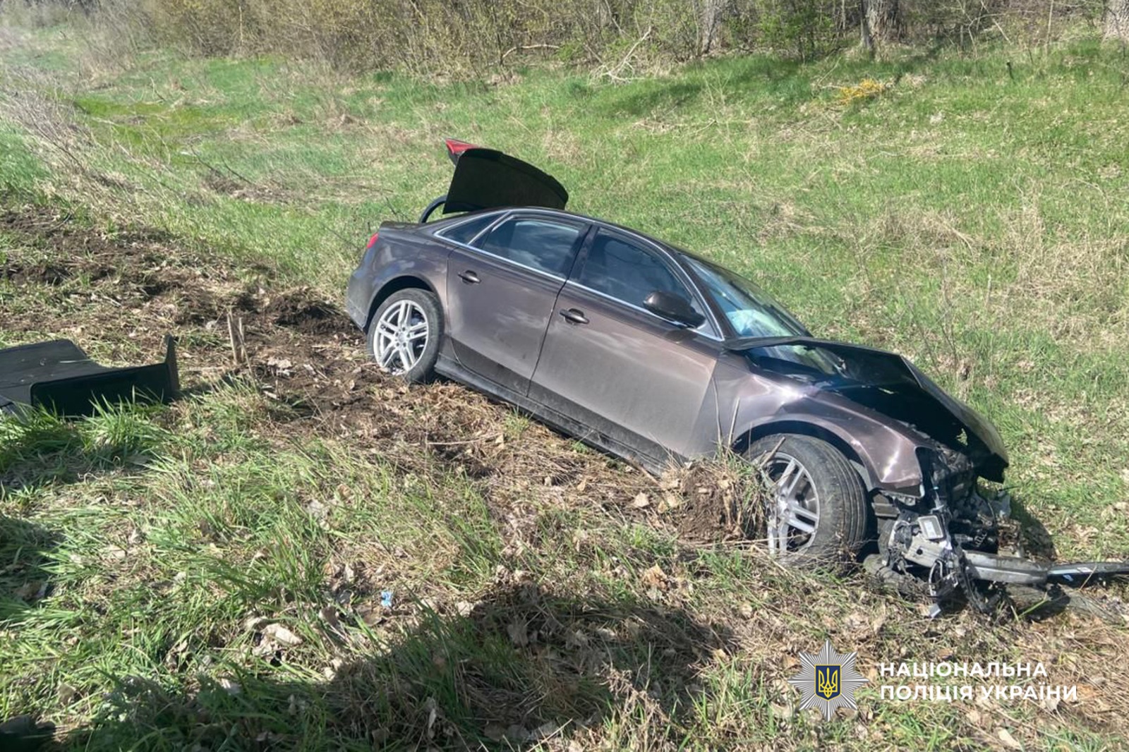 Brown sedan in a grassy ditch with heavy front-end damage and its trunk raised, полиция Ukraine watermark visible indicating a crash scene.