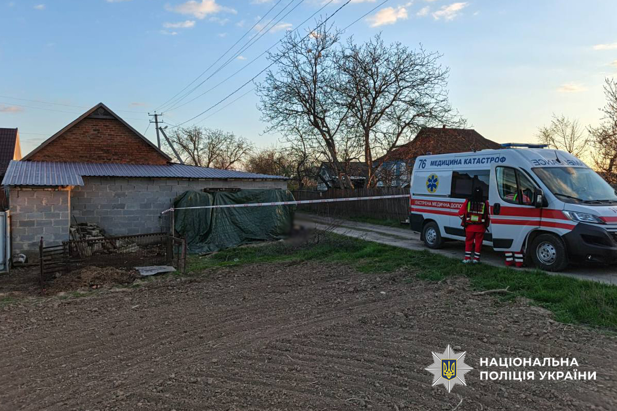 Ambulance and two medics in red uniforms on a dirt road beside a taped-off yard with a brick outbuilding in a rural area.