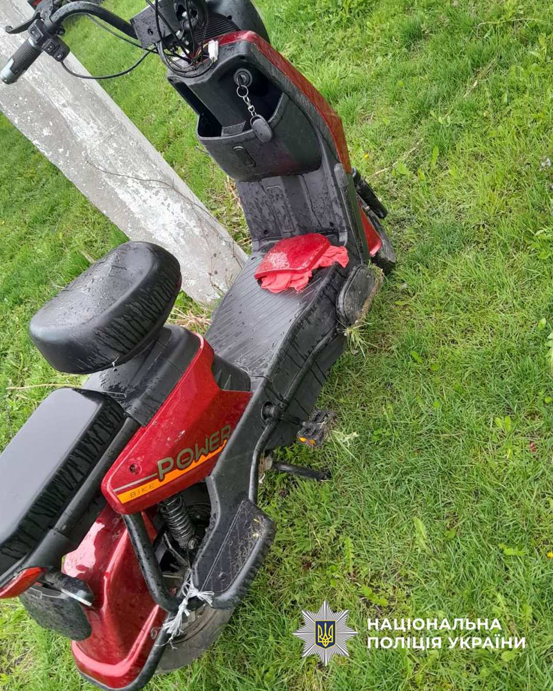Red scooter lying on its side in a grassy area, with a red cloth on the seat and a police watermark in the corner.