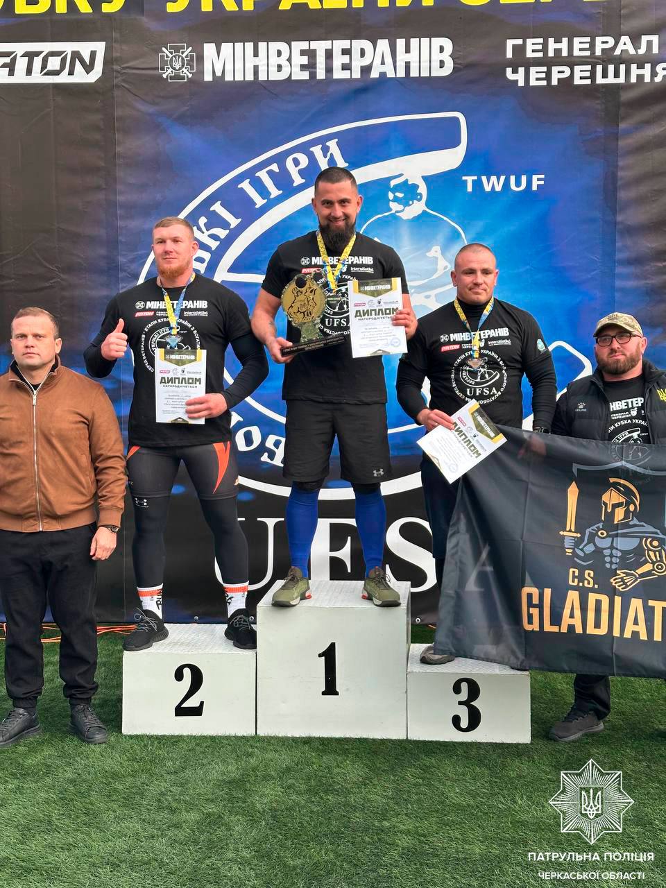 Three male competitors stand on a podium—1st in the center with a trophy and diploma, 2nd on the left, 3rd on the right with diplomas—holding a banner behind them.