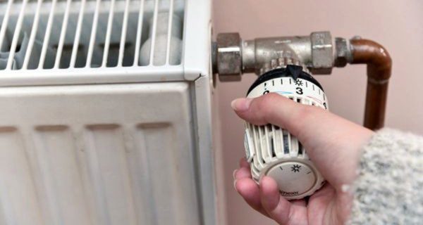 Close-up of a hand adjusting a radiator temperature dial on a metal radiator with copper piping nearby.