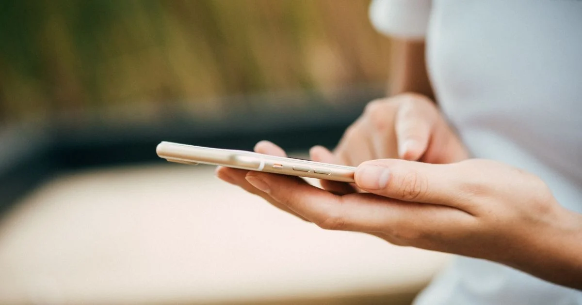 Person's hands holding a beige smartphone, fingers on the screen outdoors with a blurry background.