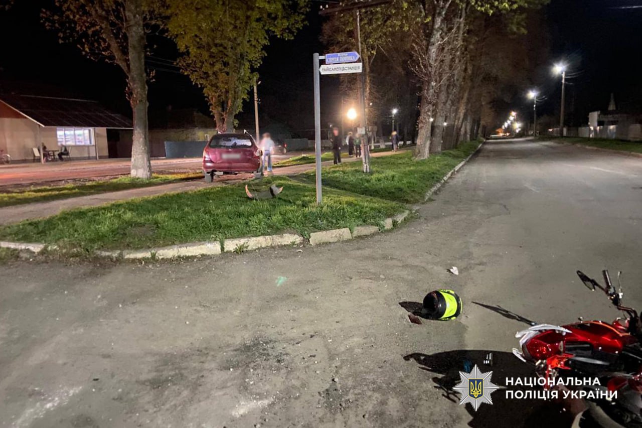 Nighttime road scene of a motorcycle crash: a damaged red motorcycle on the street, a helmet and debris on the ground, a car parked on the curb, and police presence in the distance with Ukrainian police insignia visible.