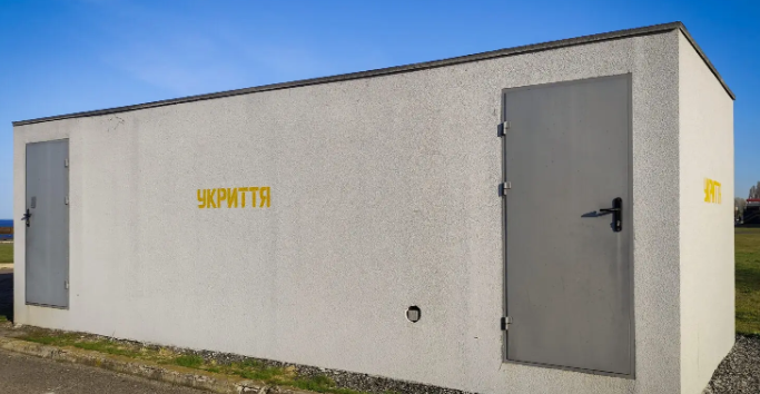 Small concrete shelter with two gray doors under a blue sky; yellow ‘УКРИТТЯ’ text on the side wall.