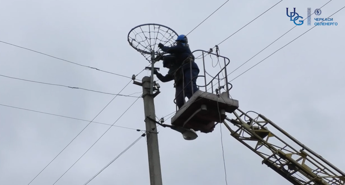 Utility worker in a blue helmet on a bucket lift repairing a streetlight on a power pole with overhead lines in a cloudy sky