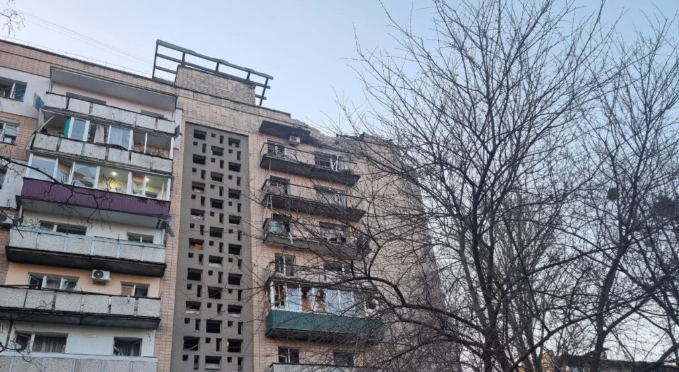 Six-story apartment building with balconies and a decorative central wall, leafless trees in the foreground.