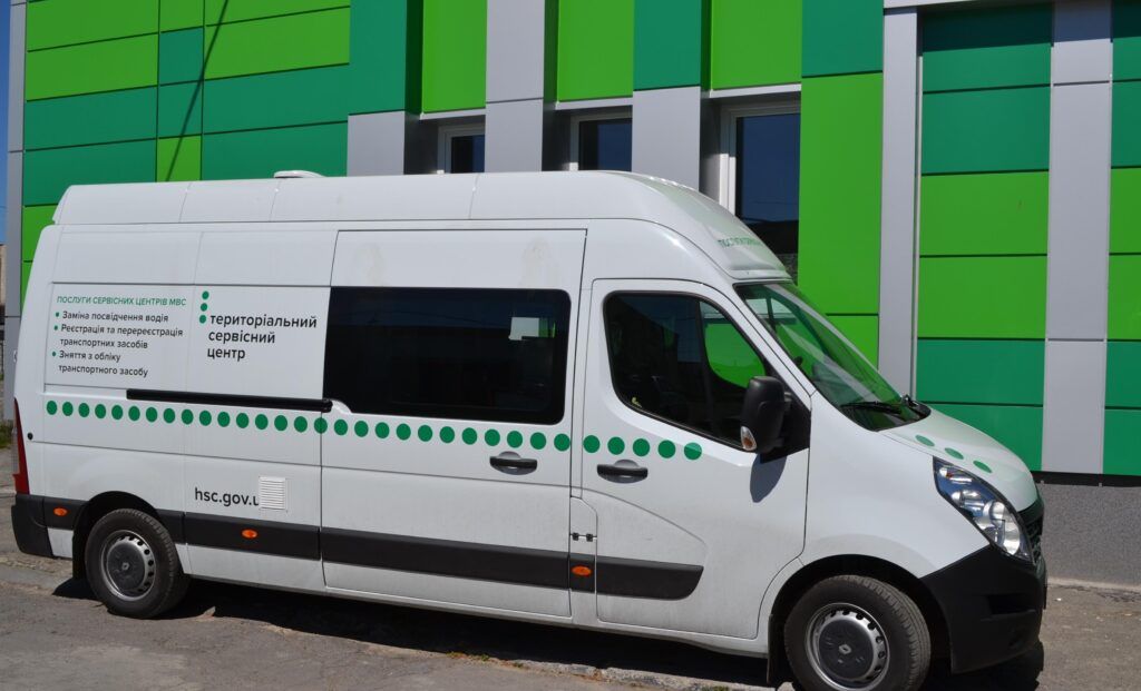White service van with green dots and Ukrainian text, in front of a green-panel building.