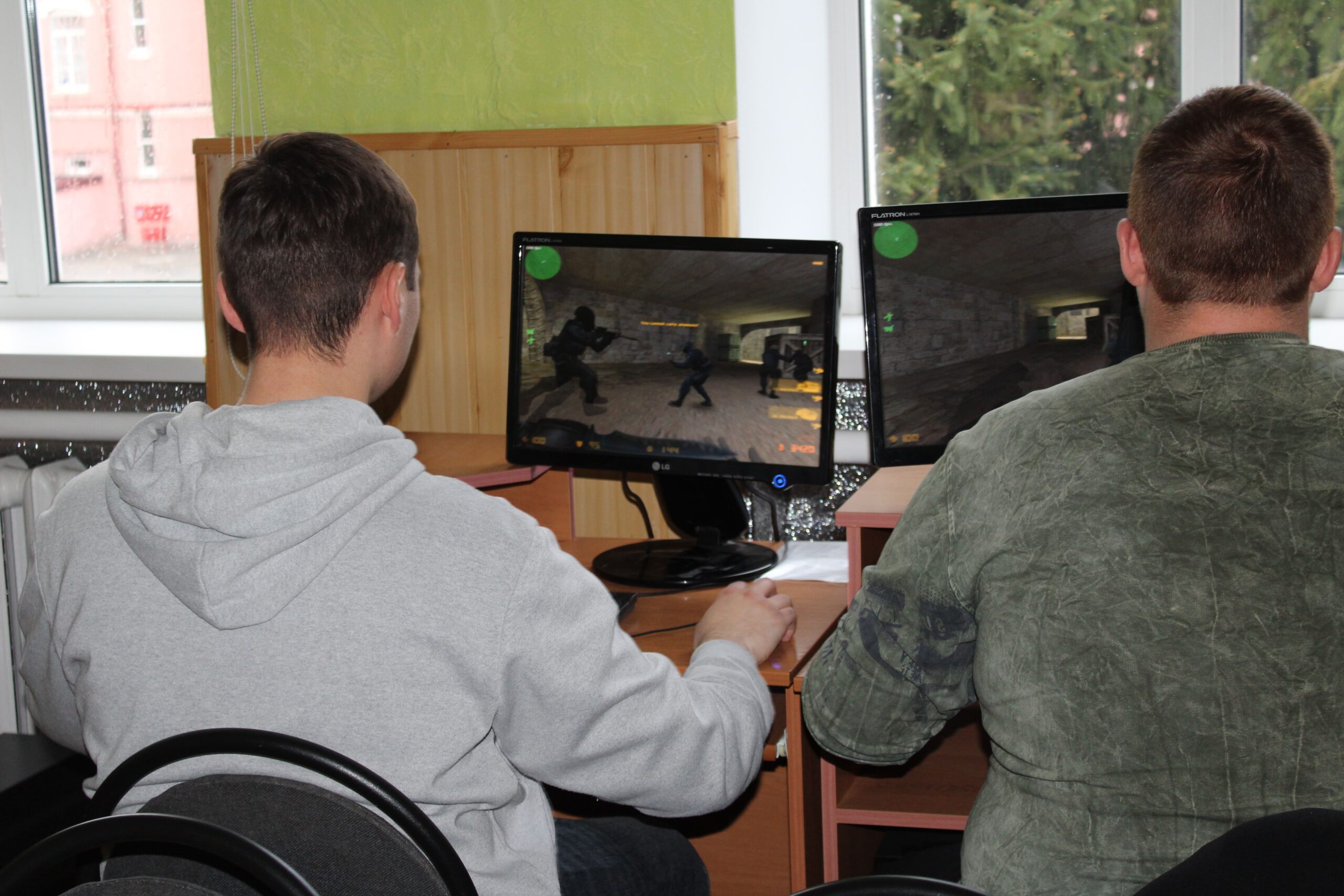 Two men sit with their backs to the camera, playing a first-person shooter on dual computer screens in a room.