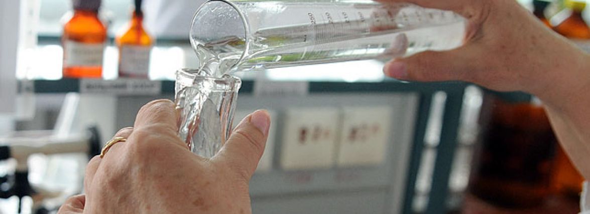 Hands pour a clear liquid from a beaker into a glass in a lab setting with orange bottles in the background