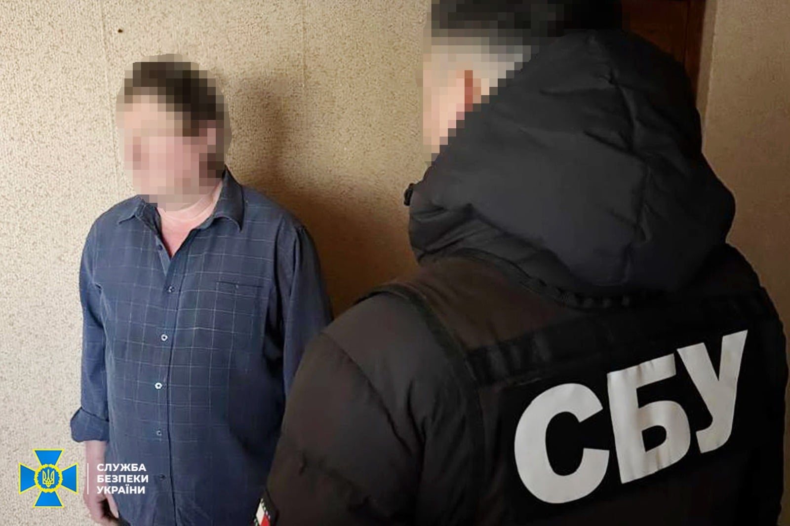 A man in a blue checked shirt stands against a beige wall while a second person in a black jacket with large white Cyrillic letters 'СБУ' stands nearby; faces are blurred.