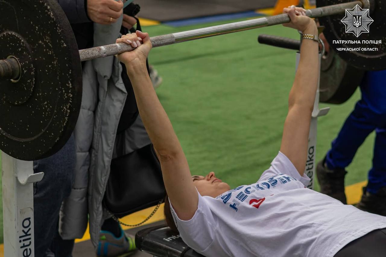 Person lying on a bench, lifting a barbell with spotters nearby; Ukrainian police logo visible in corner, gym mats underfoot.