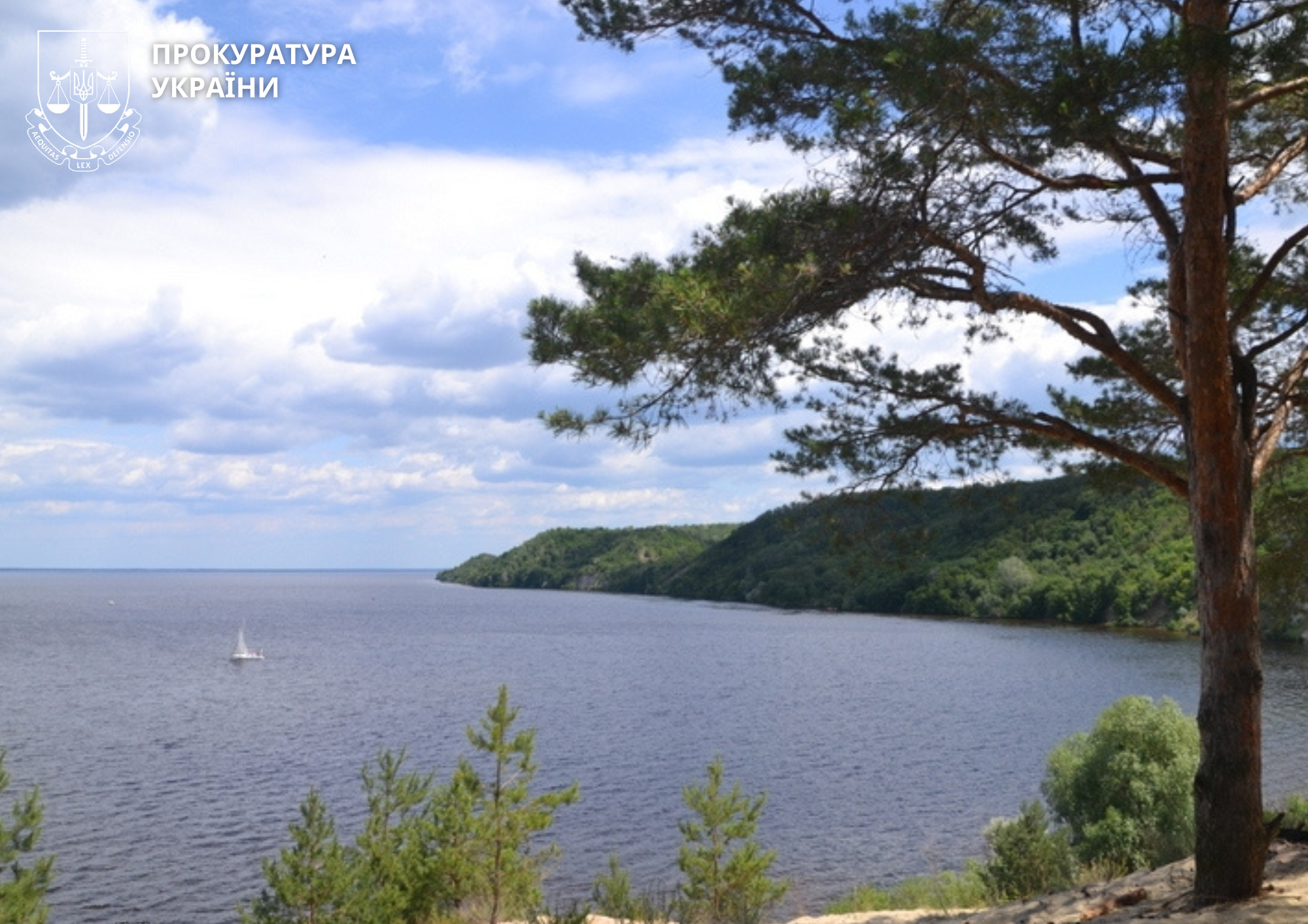 Lakeside scene with a small sailboat on calm water, tree-lined shore and cloudy sky in the background.