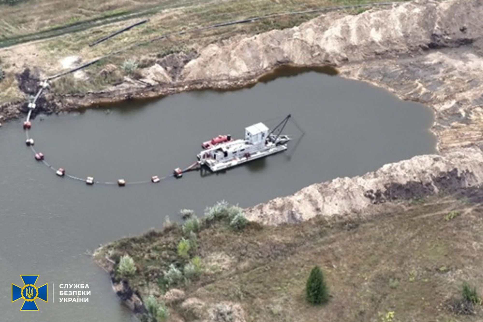 Dredging barge anchored in a circular quarry pond, connected to a buoyed hose line along the water edge. Logo of the Ukrainian security service appears in the bottom-left corner.