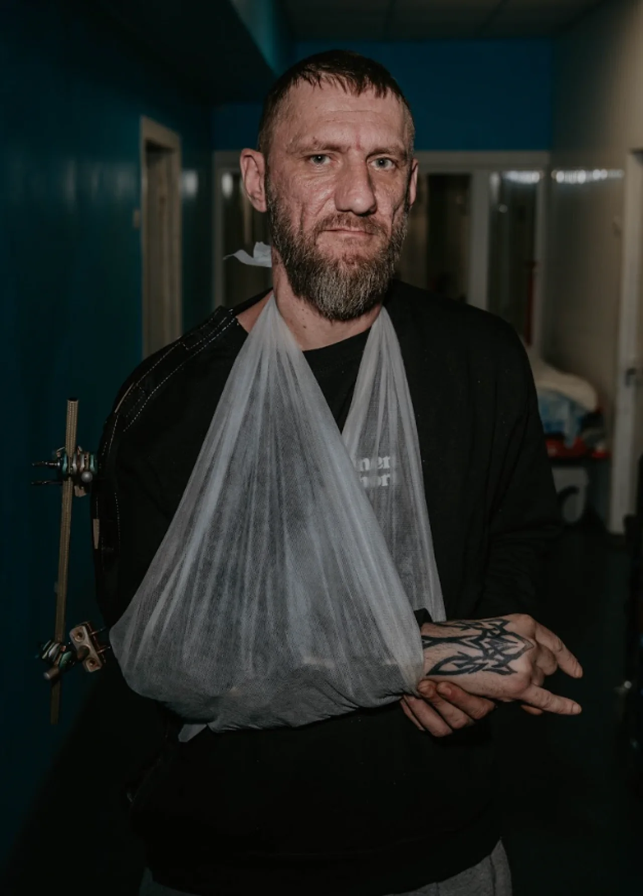 Bearded man in a black shirt wearing a gray arm sling, standing in a dim hospital corridor.