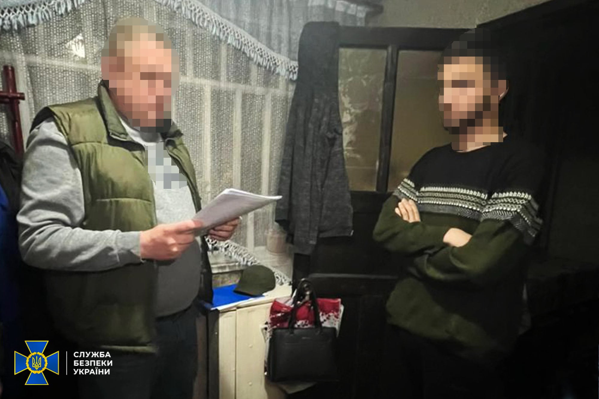 Two men indoors; one reads a paper while the other stands with arms crossed, in a room with a Security Service of Ukraine emblem in the corner.