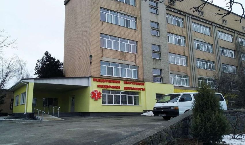 Multi-story beige brick hospital with a yellow single-story entrance and red Cyrillic signage; a white van is parked in front by the entrance.