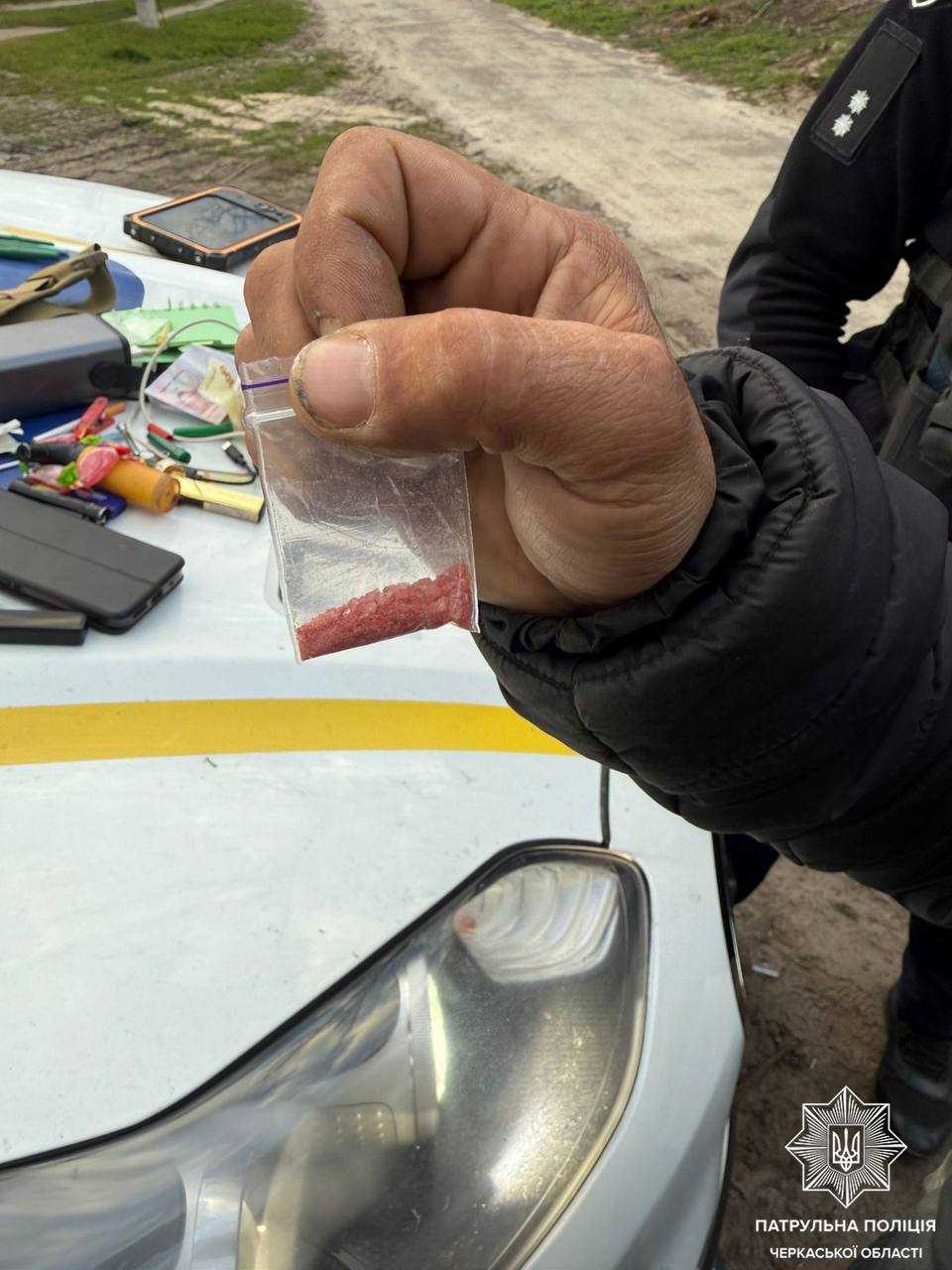 Police officer showing a small bag with red powder (drugs) on the hood of a patrol car; Ukrainian police visible in background.