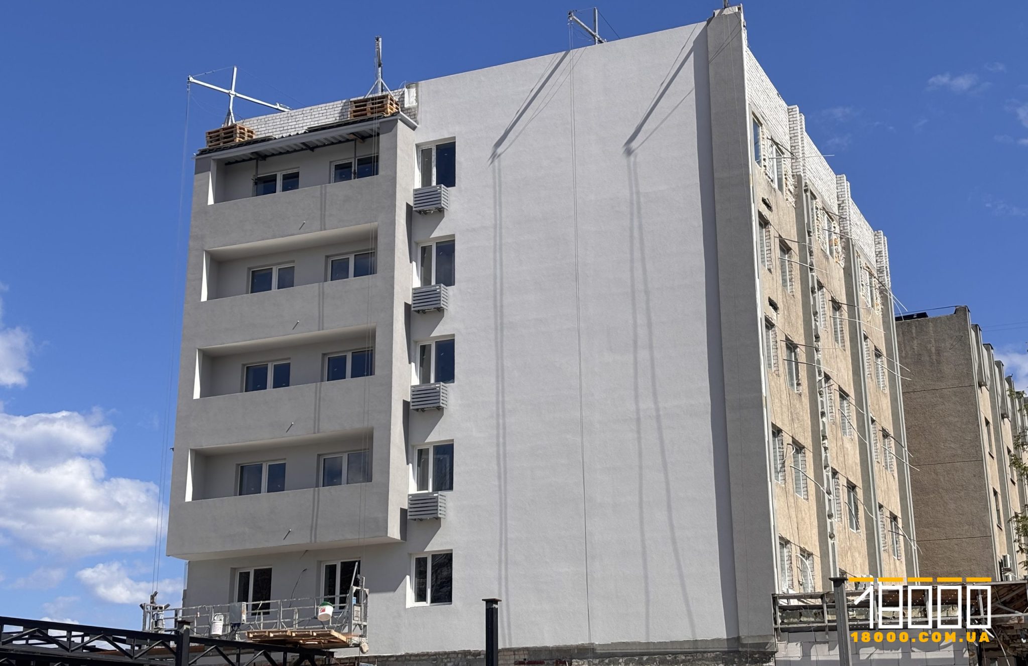 Modern gray apartment building under construction, with balconies on the left and an unfinished brick building on the right, under a clear blue sky.