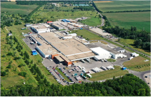 Aerial view of a large manufacturing campus with beige warehouse buildings, a parking lot full of cars, and surrounding green fields.