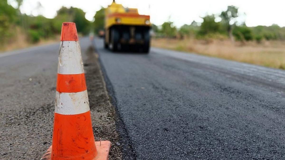 Orange traffic cone on a paved road with a road roller in the distance during asphalt paving work