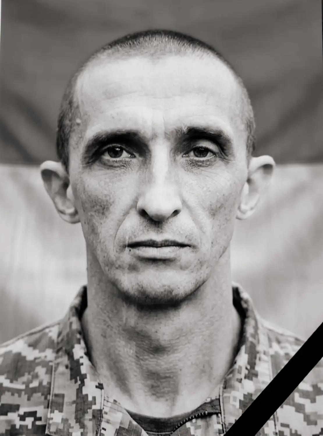 Black-and-white portrait of a man in camouflage uniform, looking straight ahead, with a diagonal black mourning ribbon across his chest.