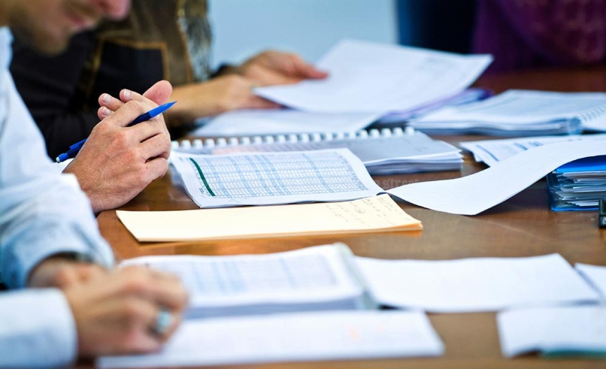 Close-up of hands writing with a blue pen among scattered papers, notebooks, and folders on a wooden conference table.