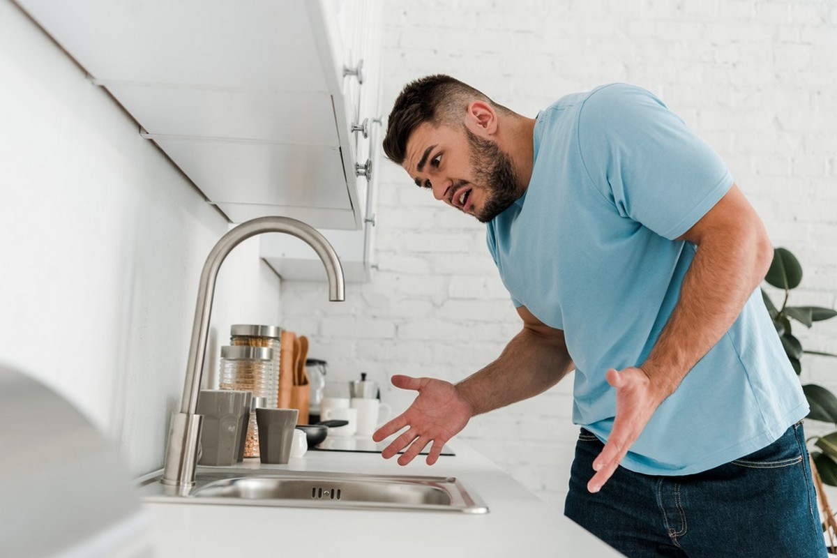 Man in a light blue shirt leans toward a modern kitchen sink, looking puzzled at the counter.