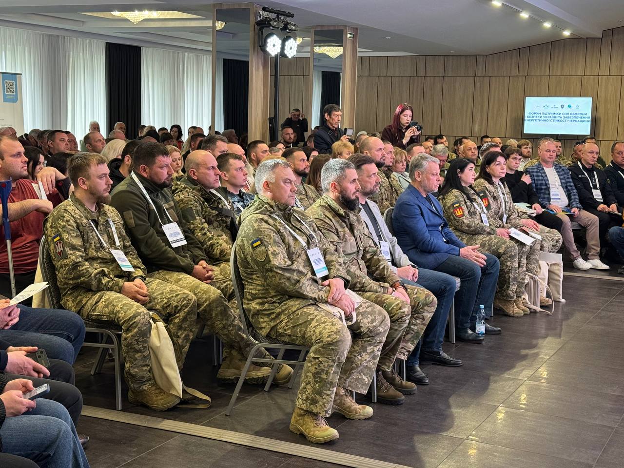 Audience at a conference: front row in camouflage uniforms, others in civilian clothing, seated in a well-lit hall with a screen and lights overhead.