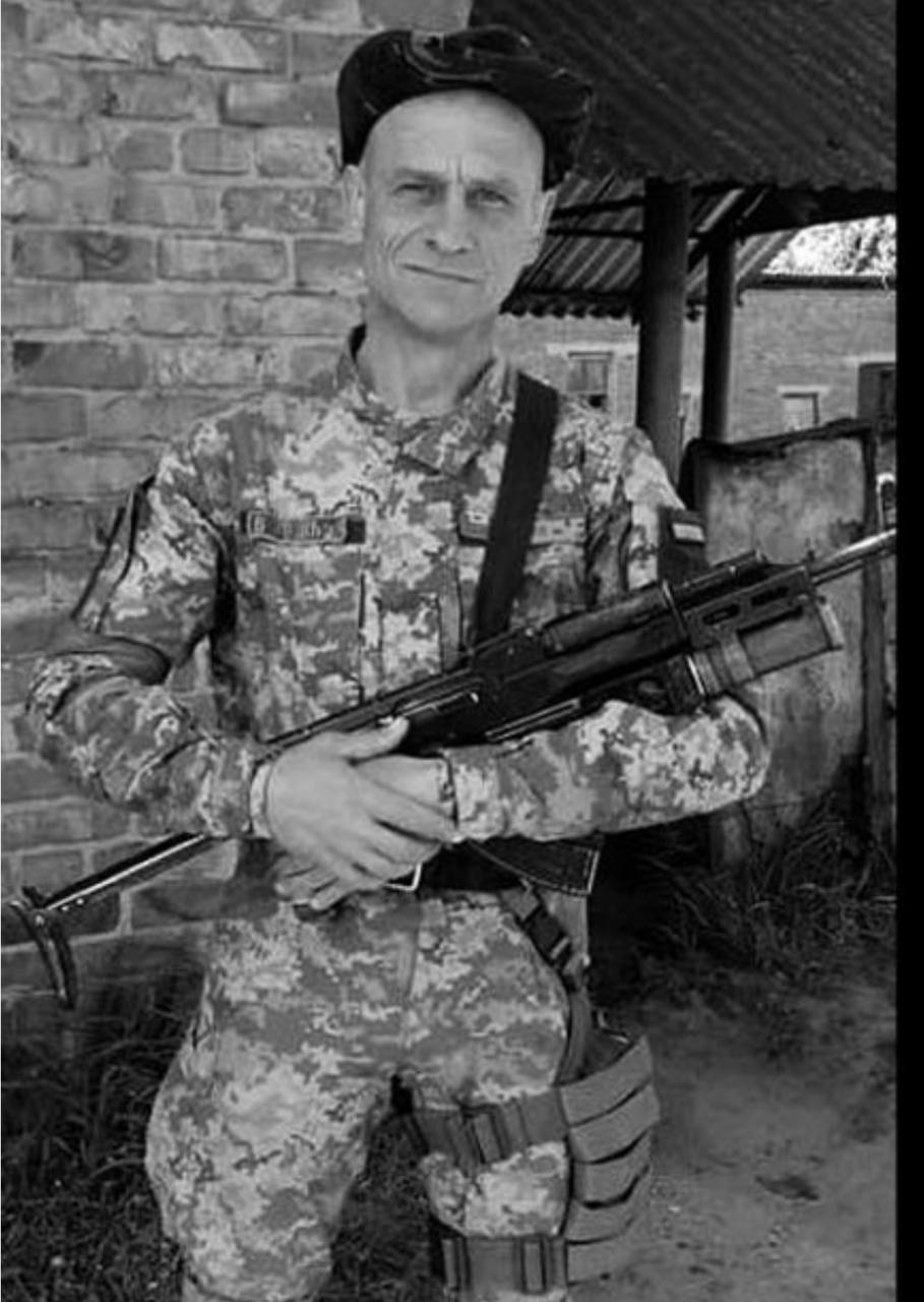 Soldier in camouflage holding a rifle across his chest, standing beside a brick wall.