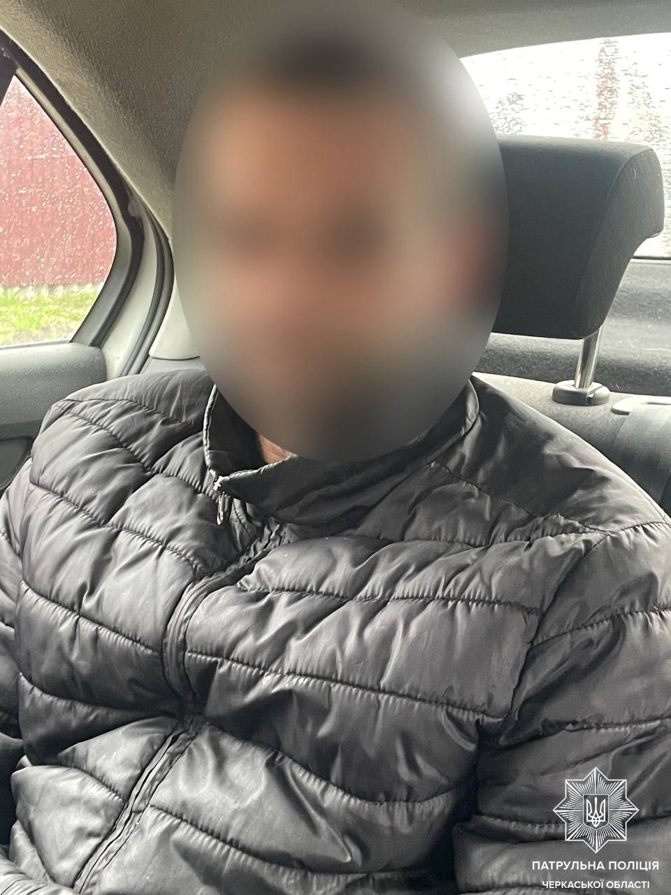 Person seated in a car wearing a dark puffer jacket; their face is blurred for privacy, with a police emblem in the bottom right corner.