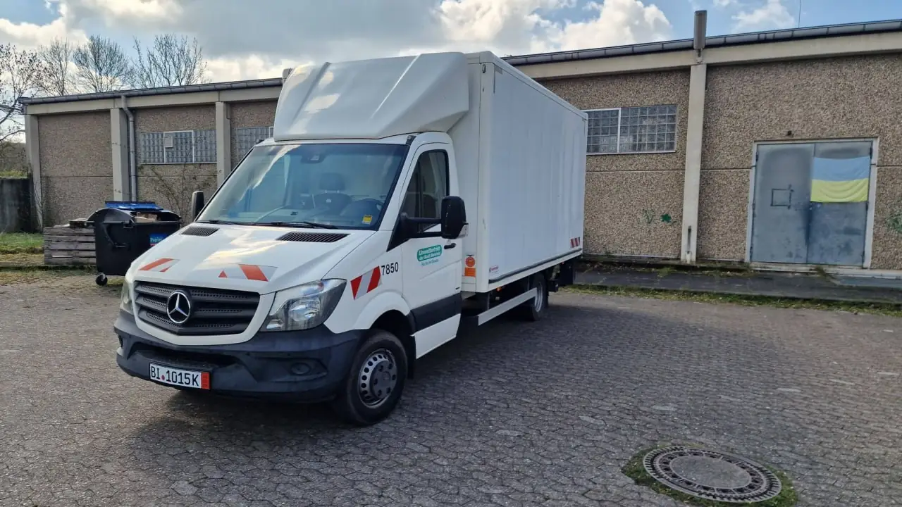 White Mercedes box truck with orange chevron safety markings parked in an industrial yard, front license plate BL 1015K, large white cargo area behind cab.
