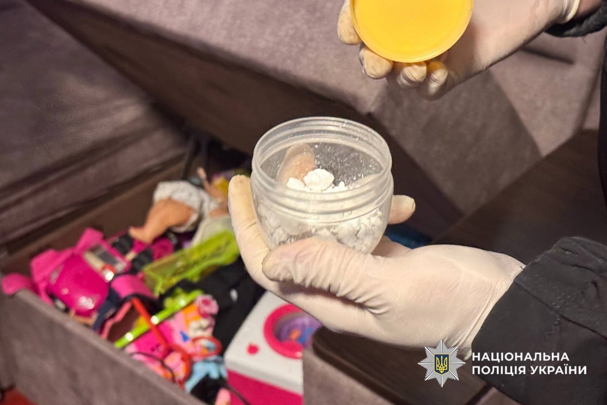 Gloved hands hold a clear jar with white powdery substance; cluttered items in the background, police evidence scene (Ukraine Police watermark).