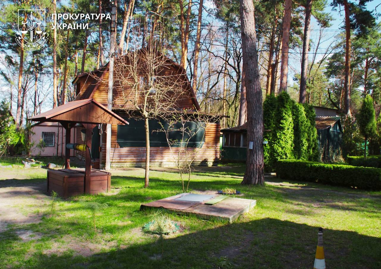 Wooden cabin in a pine forest with a small covered well in front and a grassy yard; Ukrainian Prosecutor's Office logo in the top-left corner.