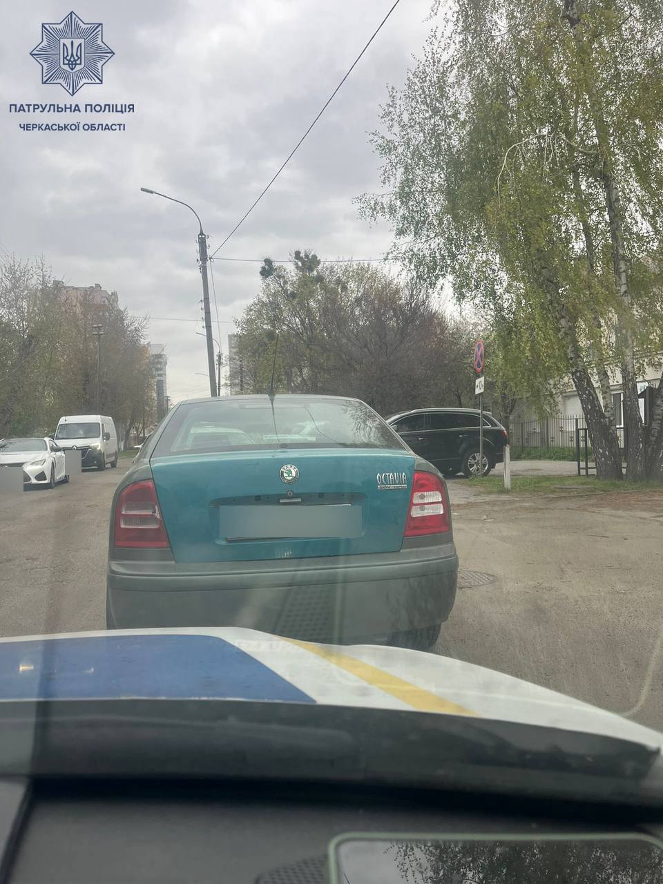 Rear view of a teal Skoda Octavia ahead on a city street, with trees on the right and other parked cars; police watermark in top-left.
