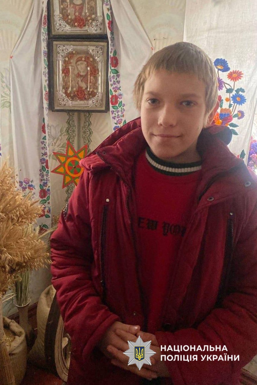 Young boy in a red jacket stands indoors in a room decorated with embroidered textiles and religious icons on the wall; Ukrainian police watermark in the bottom corner.