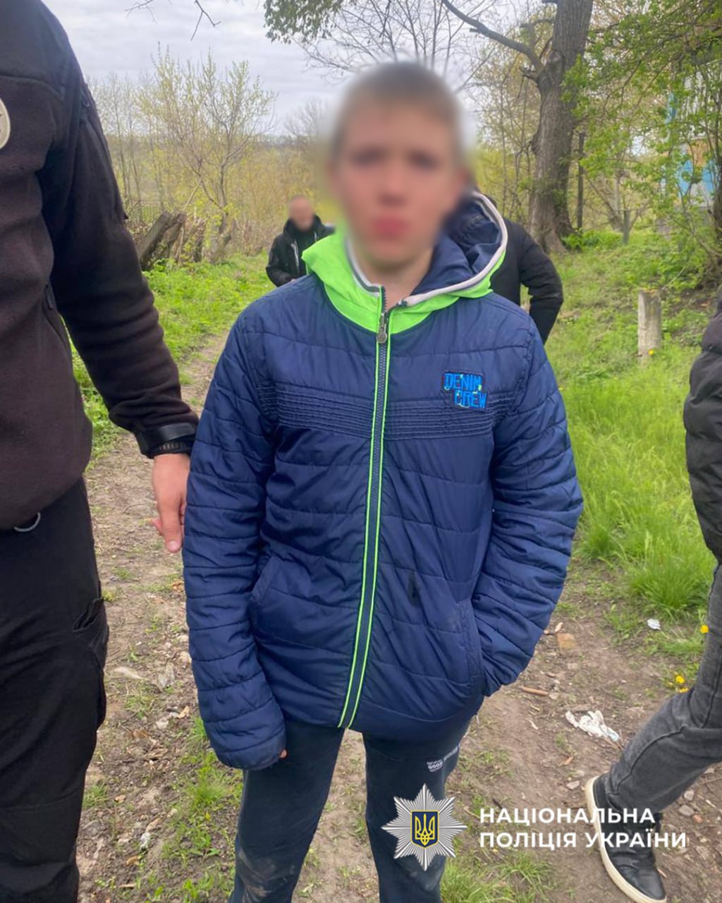 Young person in a blue puffer jacket with a neon green zipper stands on a dirt path, others nearby; Ukrainian National Police watermark visible.