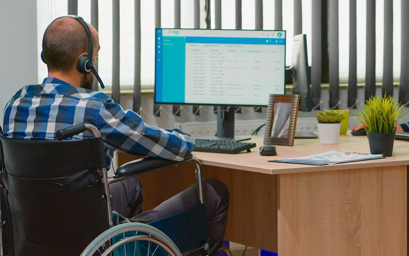 Man in a wheelchair wearing a headset sits at a wooden desk, working on a computer monitor with keyboard and papers nearby.”,”Accessibility-themed office scene: person in a wheelchair using a computer with headphones at a desk.”,”Person with a mobility impairment working at a desk with a computer monitor, keyboard, and plants nearby.”,”Wheelchair user on a call or video conference, seated at a desk with a single monitor and office setup.”,”A calm workspace showing a man in a wheelchair typing at a desk with monitor, plant decor, and papers.