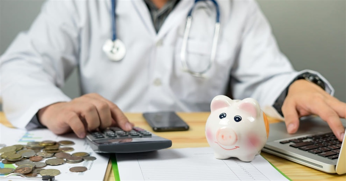 Doctor in a white coat using a calculator at a desk with a piggy bank, coins, a laptop, and financial papers nearby.