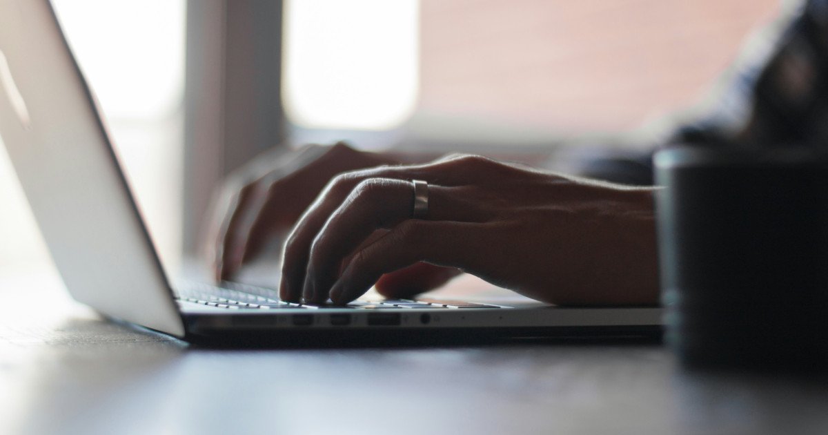 Person typing on a laptop keyboard; close-up of hands with a ring on a desk.
