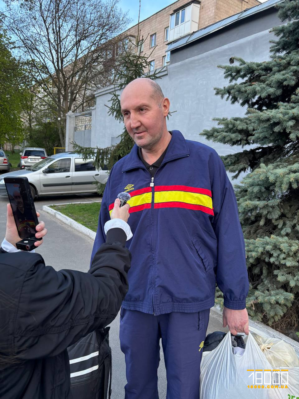 Man in a blue uniform with red and yellow stripes being interviewed outdoors, microphone near his chest; he’s holding plastic bags.