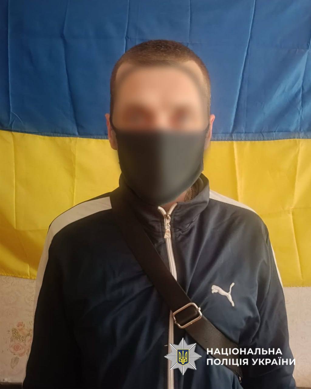 Person wearing a gray mask and dark jacket with a crossbody bag, standing before a blue-yellow Ukrainian flag; police logo bottom right.