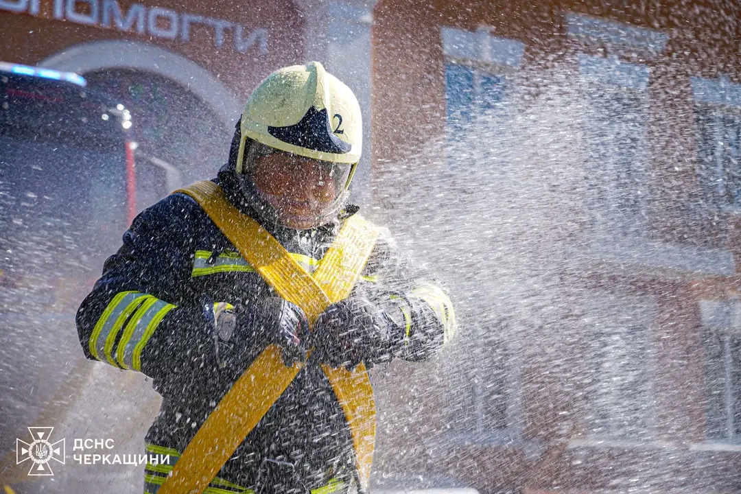 Firefighter in full turnout gear and helmet handling a bright yellow hose as water sprays around him.