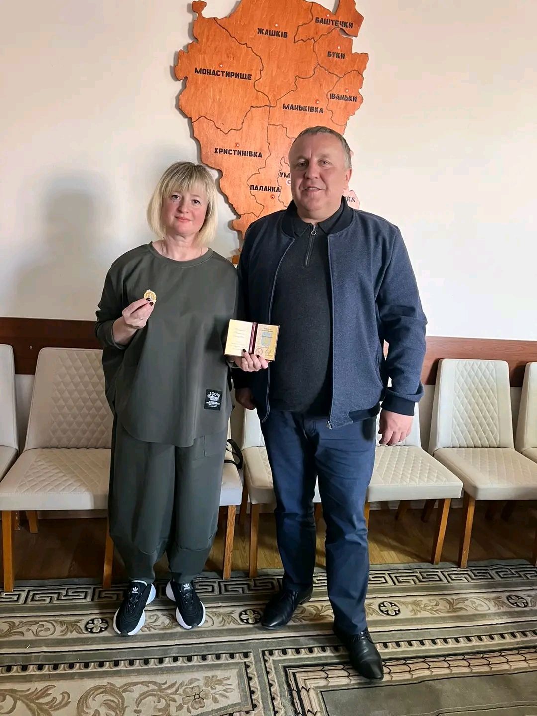 Two adults pose indoors after an award: a woman in green holds a medal; a man in a dark jacket holds a small box, with a carved map on the wall behind them.
