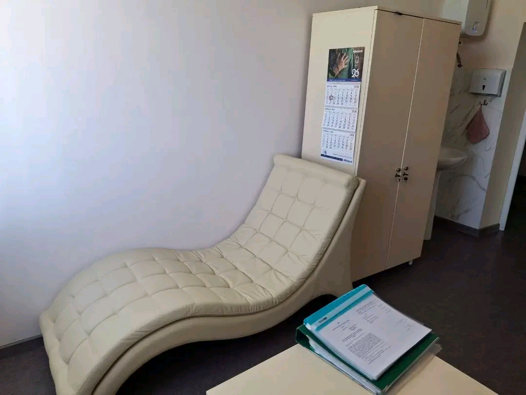 Cream padded chaise lounge with tufted upholstery in a small medical or wellness room, next to a tall beige cabinet and a calendar posted on its side.