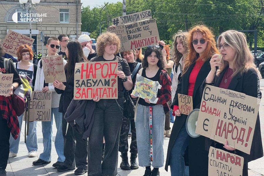 A group of protesters outdoors, holding handmade signs with Ukrainian text and red letters, addressing political demands.