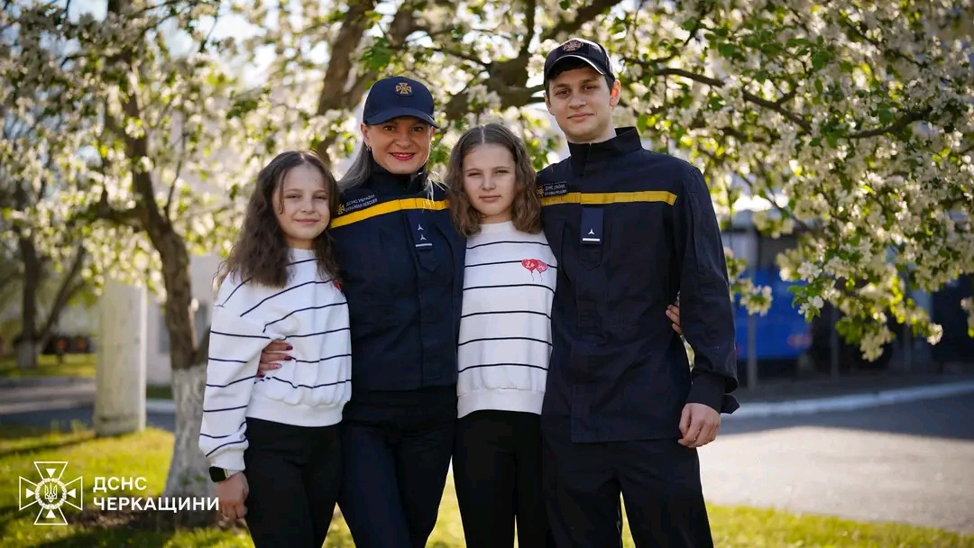 Family of four outdoors under blooming trees; mother and father in dark uniforms with yellow stripes, two girls in striped shirts.