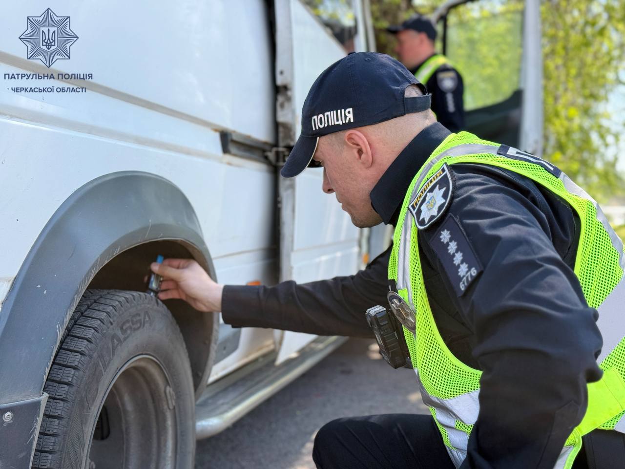 Ukrainian police officer in a neon vest kneels to check a van's front tire, using a gauge beside the wheel; patrol police logo on the vehicle wall nearby.