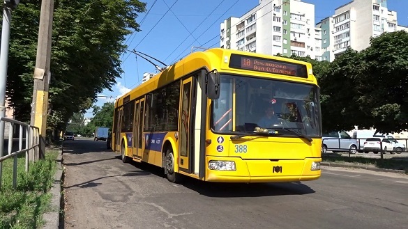 Bright yellow trolleybus driving along a city street with trees, parked cars, and apartment blocks in the background.