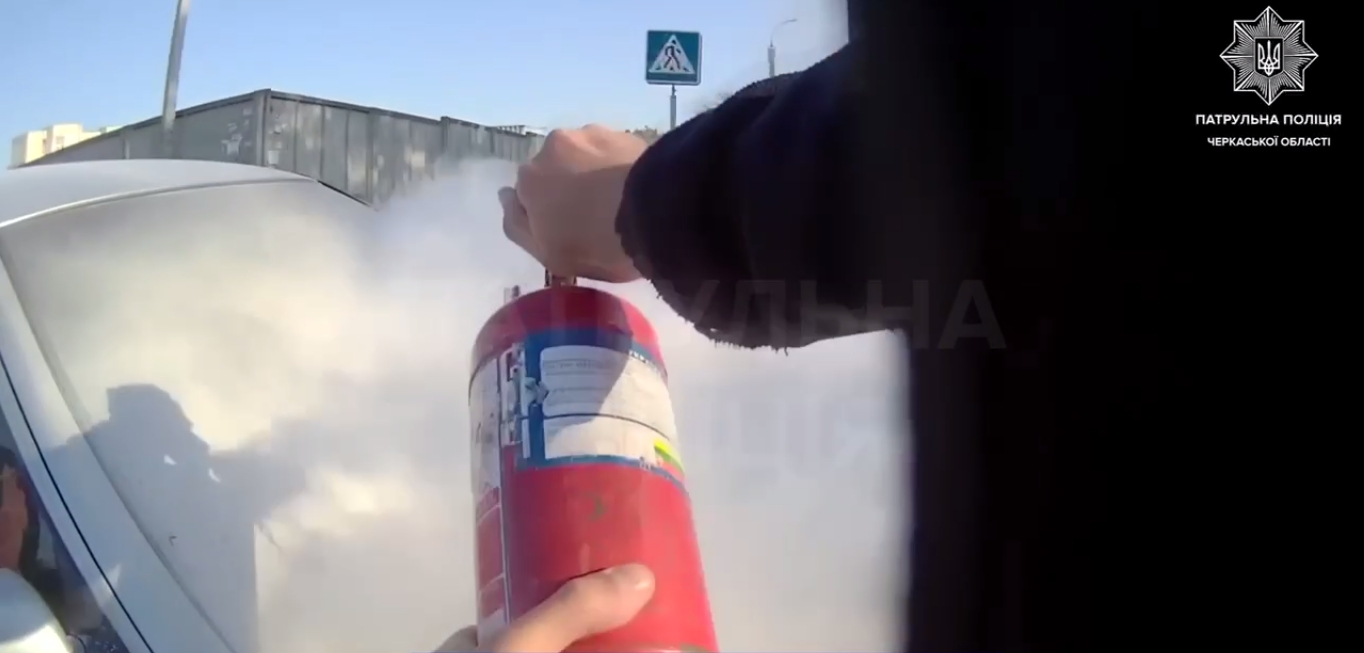 Person uses a red fire extinguisher on a car, white smoke billowing from the scene, outdoors with a pedestrian crossing sign in the background.